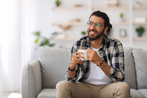 Handsome young arab man with tea mug looking at copy space and smiling, sitting on sofa at home, happy middle-eastern guy in casual relaxing alone during weekend, drinking coffee