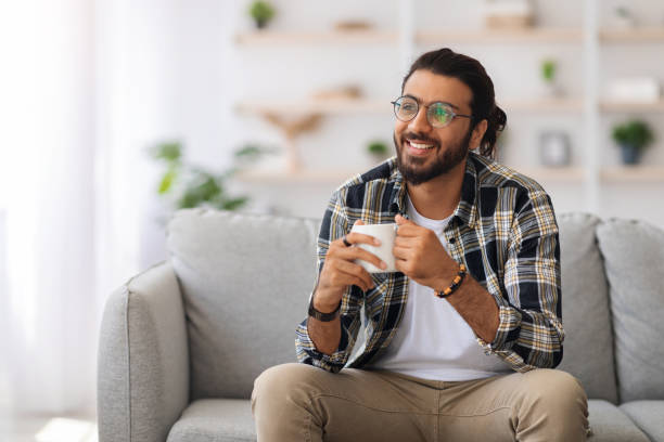 Handsome arab man with tea mug looking at copy space Handsome young arab man with tea mug looking at copy space and smiling, sitting on sofa at home, happy middle-eastern guy in casual relaxing alone during weekend, drinking coffee hot handsome man stock pictures, royalty-free photos & images