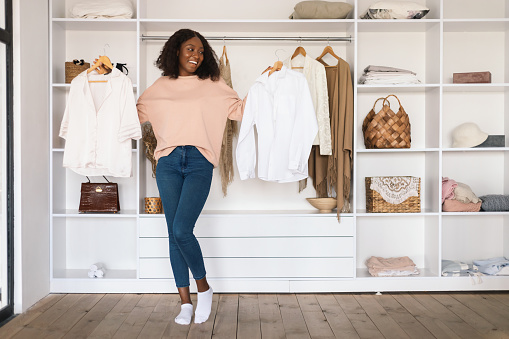 African American Female Choosing Clothes Holding Two Shirts Posing Standing In Wardrobe Room Full Of Trendy Outfits At Home. Shopaholic's Choice, Fashion And Style Concept. Full Length
