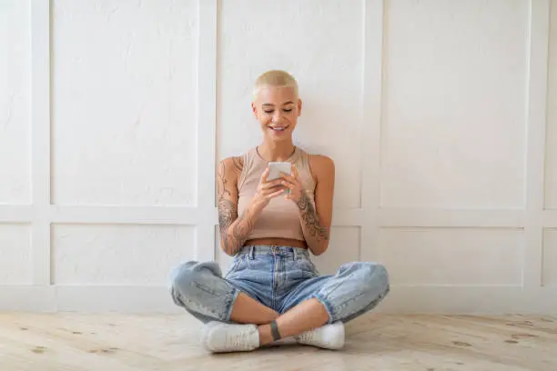 Happy lady using smartphone texting and browsing internet, sitting on floor over white wall, copy space. Casual woman networking in social media or using new app.