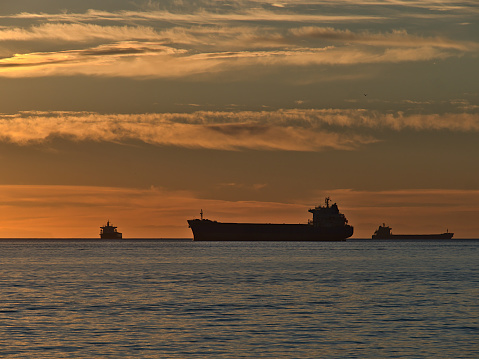 Silhouettes of three cargo freight vessels mooring in English Bay in front of Vancouver, British Columbia, Canada at sunset with orange colored dramatic sky in autumn season. Focus on ship in center.
