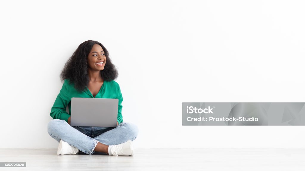 Joyful african american young woman with laptop on white Joyful african american young woman with bushy hair using laptop on white studio background, sitting on floor and looking at copy space for remote job or dating website advert, panorama Women Stock Photo