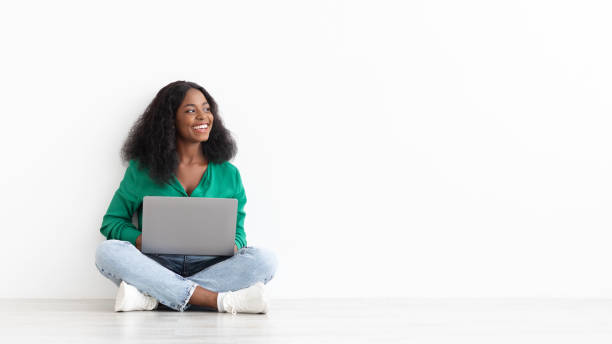 Joyful african american young woman with laptop on white Joyful african american young woman with bushy hair using laptop on white studio background, sitting on floor and looking at copy space for remote job or dating website advert, panorama online training courses stock pictures, royalty-free photos & images
