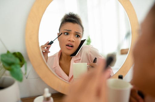 Emotional african american lady in pajamas talking on cellphone with girlfriend while putting mascara in front of mirror at home in the morning, sharing rumors