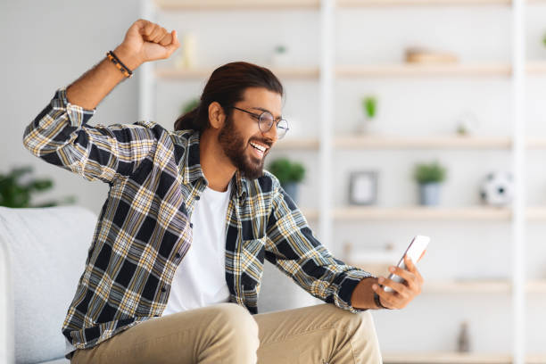 istock Emotional long-haired indian guy hipster sitting on couch, using smartphone 1352727099