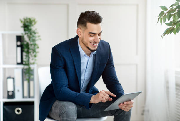 istock Cheerful young Arab businessman in formal suit using tablet computer, working online in modern office 1352727408