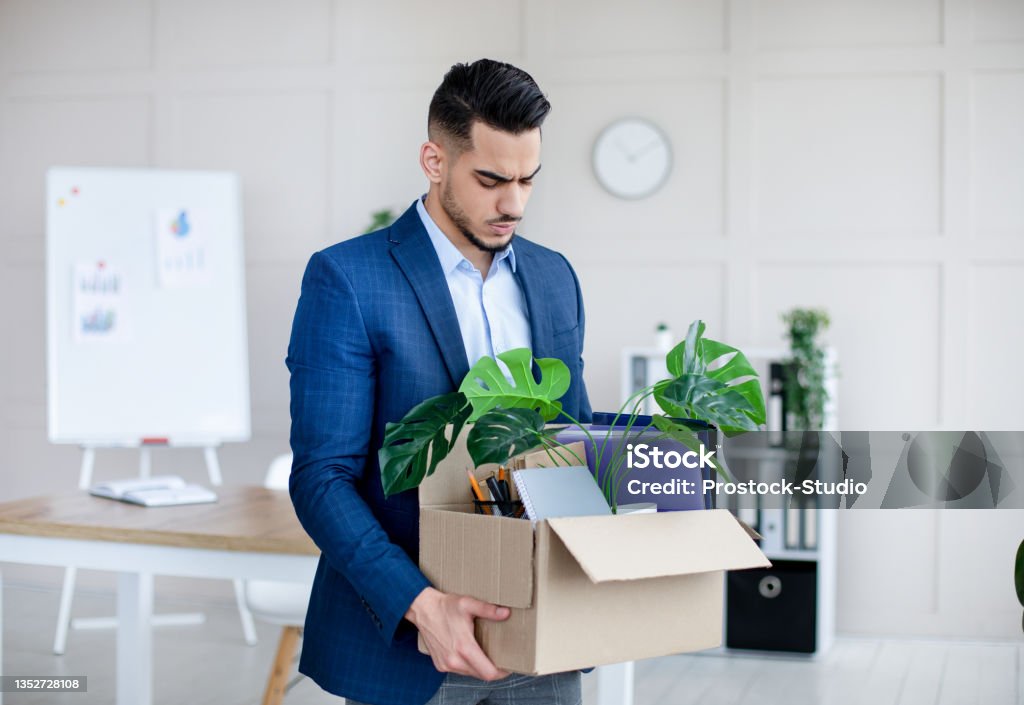 Unemployed Arab guy in formal wear holding personal belongings, feeling depressed after losing his job Unemployed Arab guy in formal wear holding personal belongings, feeling depressed after losing his job. Upset Eastern man with cardboard box of things leaving office after being fired Employee Stock Photo
