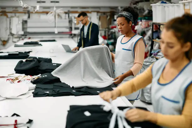 Young black seamstress and her coworkers working with fabric at clothing factory.