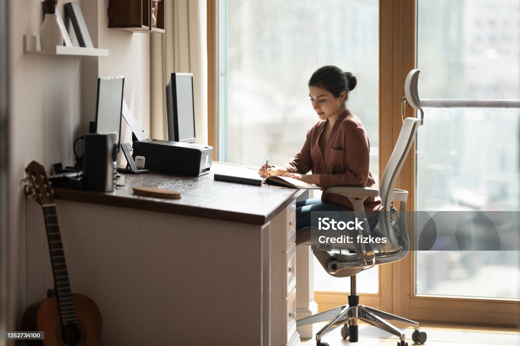 Serious Indian female sit at desk holds pen jotting information Serious young Indian female sit on ergonomic comfy chair at desk holds pen jotting information, student studying, business woman makes useful notes on paper notebook. Modern office, workflow concept Ergonomics Stock Photo