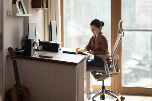 Serious Indian female sit at desk holds pen jotting information Serious young Indian female sit on ergonomic comfy chair at desk holds pen jotting information, student studying, business woman makes useful notes on paper notebook. Modern office, workflow concept home office chair stock pictures, royalty-free photos & images