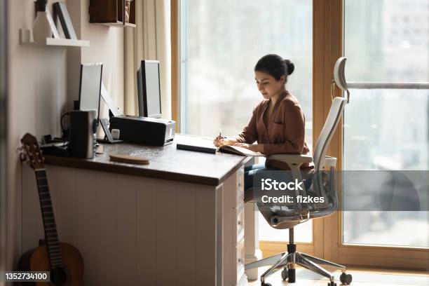 Serious Indian Female Sit At Desk Holds Pen Jotting Information Stock Photo - Download Image Now