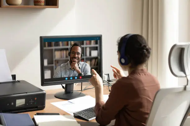 Rear back view Indian woman sit at workplace desk talk to African colleague use pc and video call app. Educational webinar participants, negotiations remotely. Online class teacher and learner concept