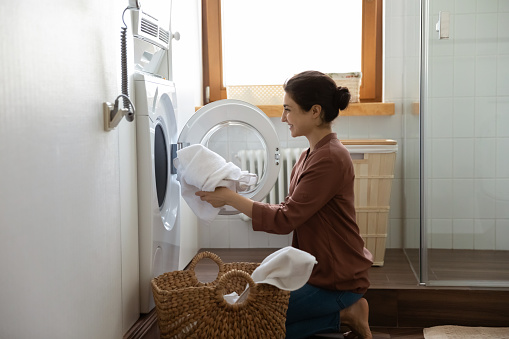 Young happy Indian housewife doing housework putting clothes into washing machine in cozy domestic laundry room. Routine house work, housekeeping, washer-dryer modern appliance or detergent ad concept