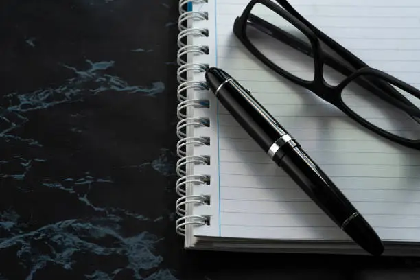 Glossy black/silver pen and matte black frame lenses on notebook striped on black background.