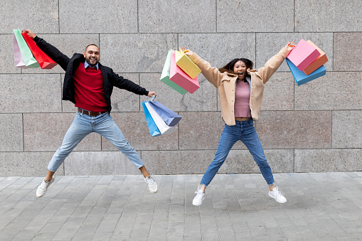 Happy young multinational couple with shopper bags jumping near supermarket, going shopping together. Positive millennial diverse family flying in air, celebrating great purchases next to mall