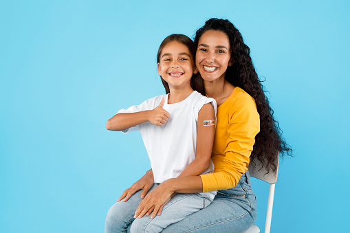 Arabic Girl And Her Mom Showing Vaccinated Arm And Thumbs Up Gesture After Covid-19 Vaccination Sitting Over Blue Background, Studio Shot. Coronavirus Protection For Kids Concept