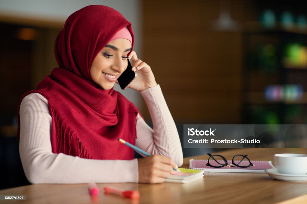 Beautiful muslim woman talking on cellphone at cafe Beautiful young muslim woman in hijab talking on cellphone at cafe, drinking coffee and taking notes. Cheerful middle-eastern lady looking for job, having conversation with employer, copy space Interview - Event Stock Photo