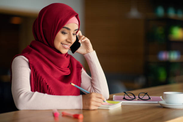 Beautiful muslim woman talking on cellphone at cafe Beautiful young muslim woman in hijab talking on cellphone at cafe, drinking coffee and taking notes. Cheerful middle-eastern lady looking for job, having conversation with employer, copy space cafe racer stock pictures, royalty-free photos & images