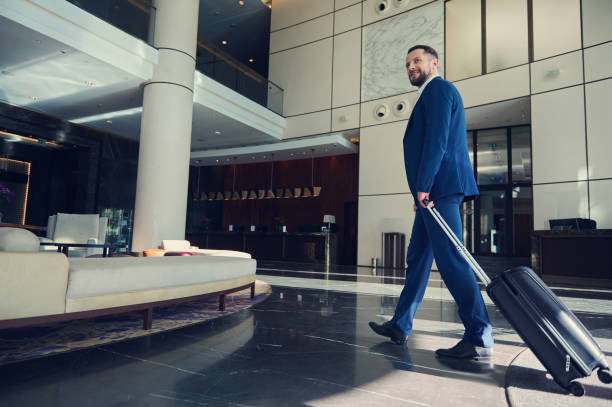 hombre guapo, exitoso desarrollador de negocios sonriendo, mirando a un lado, caminando por el vestíbulo, dirigiéndose con equipaje a la sala de estar dentro de un moderno edificio de lujo durante el viaje. - hotel reception lobby hotel luxury fotografías e imágenes de stock