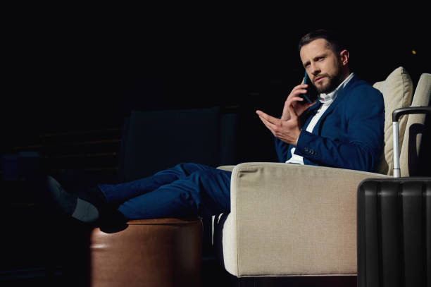 istock Confident successful and perplexed young European businessman talking on a mobile phone while sitting in an armchair of a hotel lounge, relaxing after a business trip. Suitcase near armchair 1352750304