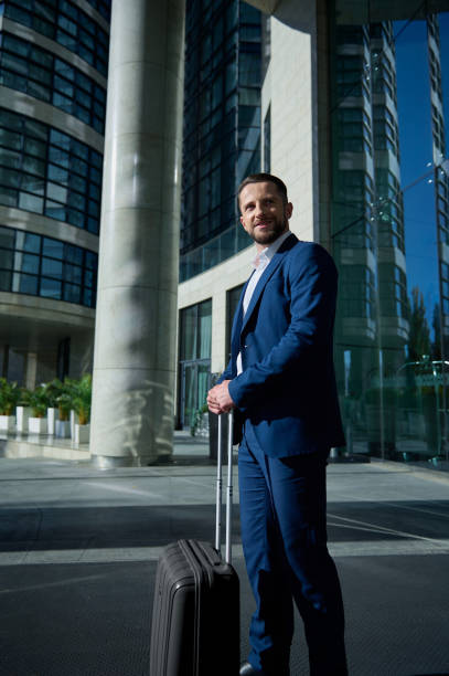 istock Self-confident attractive successful businessman developer entrepreneur in business suit and luggage smiles looking aside standing outdoor on a corporate high-rise building glass entrance background 1352750445