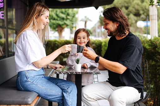 A young family came together in a cafe. Mom, dad and little daughter drink tea, eat cakes. They are happy together.