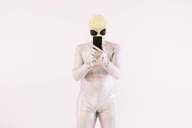 Person dressed in silver suit and green alien mask looking at mobile phone over white background stock photo
