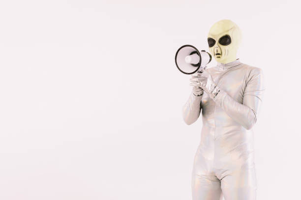 istock Person dressed in silver suit and green alien mask, speaking through a white megaphone, on white background 1352751925