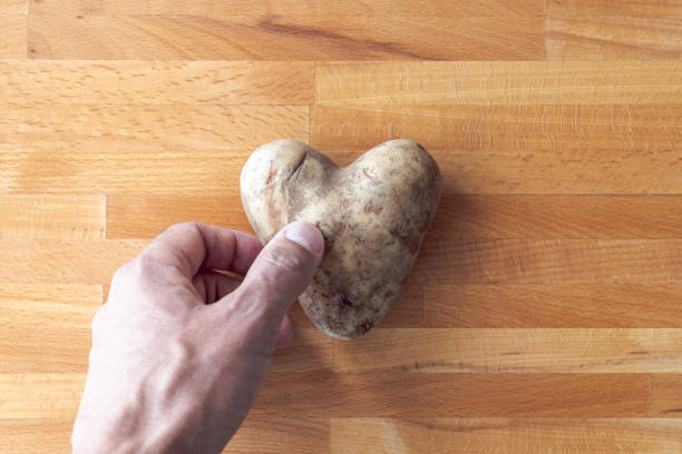 ręka mężczyzny sięga w dół, aby podnieść ziemniaka w kształcie serca leżącego na drewnianej desce do krojenia, tworząc świetne tło symbolizujące miłość do gotowania. - heart shape raw potato food individuality zdjęcia i obrazy z banku zdjęć