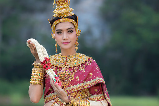 Close up of Portrait smiling young asian girl wearing thai tradition red dress and golden jewelry standing in the nature park and holding flower garlands, beautiful ancient thai dress life portrait concept