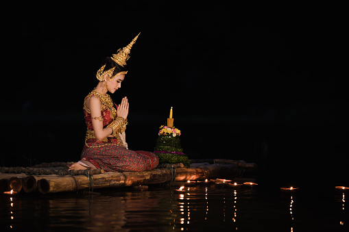 night portrait thai model wearing traditional red dress and jewelry sitting on bamboo raft at night loy kratong  festival, concept to carry on the beautiful traditions and culture of Thailand
