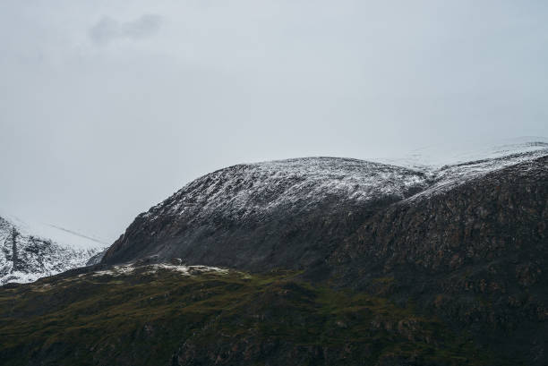 istock Minimalist highlands landscape with snow-capped mountains under cloudy sky. Snowbound high mountain range in overcast weather. Atmospheric minimalism with green mountains, white snow and gray sky. 1352766328