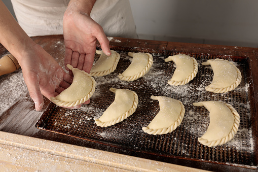 A Close up cook's hands placing raw empanadas in a baking dish on a rustic wooden table with flour. Horizontal image.