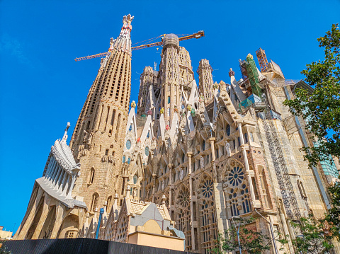 Barcelona, Spain - June 14, 2022:  Construction cranes complete the architectural details of the gothic Art Nouveau church spires of the Sagrada Familia basilica in the Eixample district of Barcelona Catalonia Spain as seen from the Gaudi Park pond