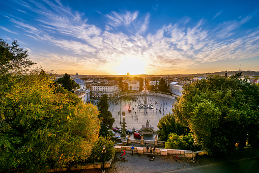 A suggestive and colorful sunset sky illuminates the roofs of Rome seen from the panoramic terrace of the Pincio Gardens, in the historic and baroque heart of the Eternal City. The terrace of the Pincio, one of the most visited and loved places in Rome, is the culmination of the west side of Villa Borghese, the public park considered the green lung in the heart of the Italian capital. In addition to offering a breathtaking 180-degree view of the city, the Pincio overlooks the famous Piazza del Popolo, an elliptical esplanade in the neoclassical style designed by the Roman architect and urban planner Giuseppe Valadier in 1816. Along the horizon the silhouette of the majestic dome of St. Peter's Basilica. In 1980 the historic center of Rome was declared a World Heritage Site by Unesco. Super wide angle and high definition image.