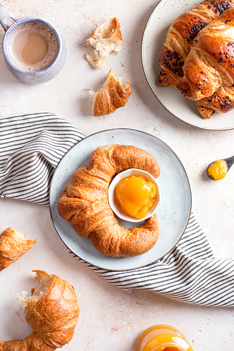 Freshly baked croissants, apricot jam and mug with cappuccino on beige stone table. French styled breakfast flat lay, top view