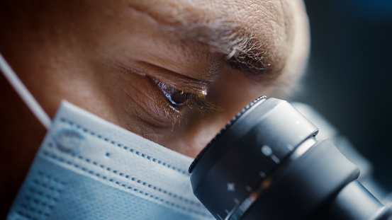 Foto de primer plano ultra macro de un científico masculino con máscara quirúrgica y mirando hacia el microscopio. Microbiólogo trabajando en muestras de moléculas en laboratorio moderno con equipos tecnológicos. photo