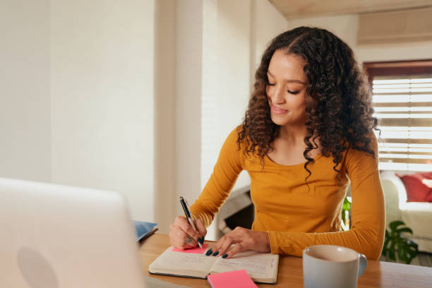 istock Happy Multi-cultural female focussed, writing in notebook. Young professional remote working with laptop in modern apartment 1352823459
