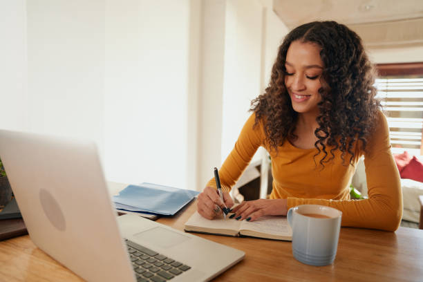 istock Multi-cultural female working from home, writing in notebook. Young professional remote working with laptop in modern apartment 1352824153