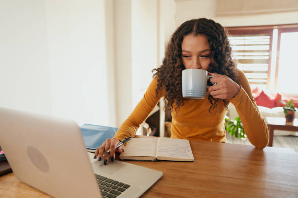 istock Multi-cultural female working from home, sipping coffee. Young professional remote working with laptop in modern apartment 1352824354