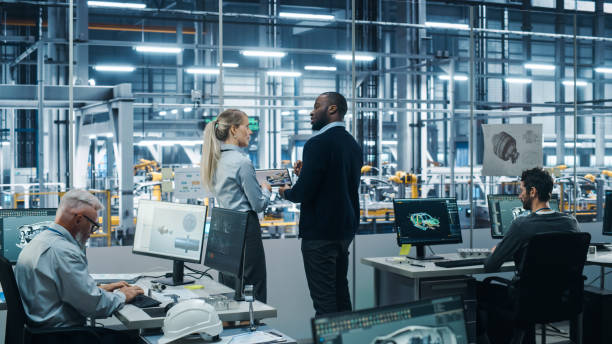 istock Car Factory Office: Team of Automotive Engineers Working, Talk, Use Tablet Computers to Design Advanced 3D Models for High-Tech Engines. Automated Robot Arm Assembly Line Manufacturing Vehicles 1352825012