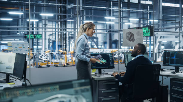 istock Car Factory Office: Female Supervisor Talks with Male Automotive Engineer, Using Computer to Design Advanced 3D Models for High-Tech Engines. Automated Robot Arm Assembly Line Manufacturing Vehicles 1352825013