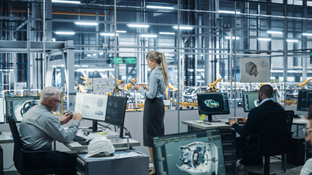 istock Car Factory Office: Team of Automotive Engineers Working, Using Computers to Design Advanced 3D Models for High-Tech Engines. Automated Robot Arm Industrial Assembly Line Manufacturing Vehicles 1352825014