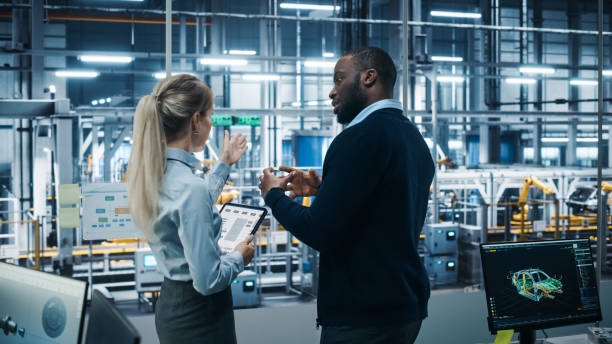 istock Car Factory Office: Female Engineer Talks with Male Scientist, Use Tablet Computer to Design Production Conveyor for Advanced Power Engines. Automated Robot Arm Assembly Line Manufacturing Vehicles 1352825019