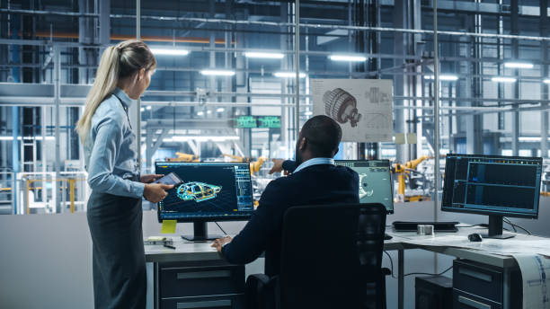 istock Car Factory: Female Supervisor Talks with Male Automotive Engineer, Using Computer to Design Advanced 3D Models for High-Tech Engines. Automated Robot Arm Assembly Line Manufacturing Electric Vehicles 1352825025