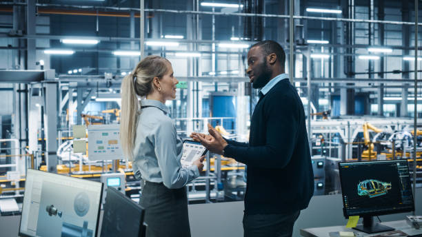 istock Car Factory Office: Female Engineer Talks with Male Scientist, Use Tablet Computer to Design Production Conveyor for Advanced Power Engines. Automated Robot Arm Assembly Line Manufacturing Vehicles 1352825027