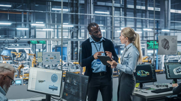 istock Car Factory Office: Female Engineer Talks with Male Scientist, Use Tablet Computer to Design Production Conveyor for Advanced Power Engines. Automated Robot Arm Assembly Line Manufacturing Vehicles 1352825029