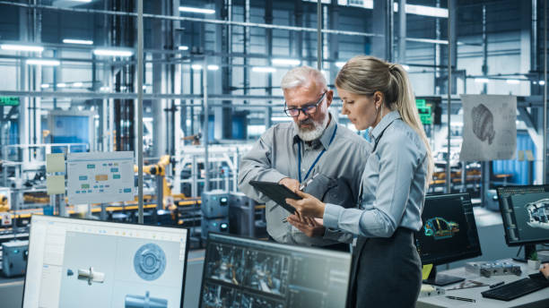 istock Car Factory Office: Manager Talks with Senior Engineer, Use Tablet Computer to Design Production Conveyor for Advanced Power Engines. Automated Robot Arm Assembly Line Manufacturing Vehicles 1352825036