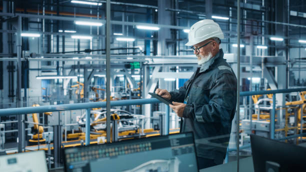 istock Car Factory: Male Automotive Engineer Wearing Hard Hat, Standing Using Tablet Computer. Monitoring, Control, Equipment Production. Automated Robot Arm Assembly Line Manufacturing Electric Vehicles 1352825037