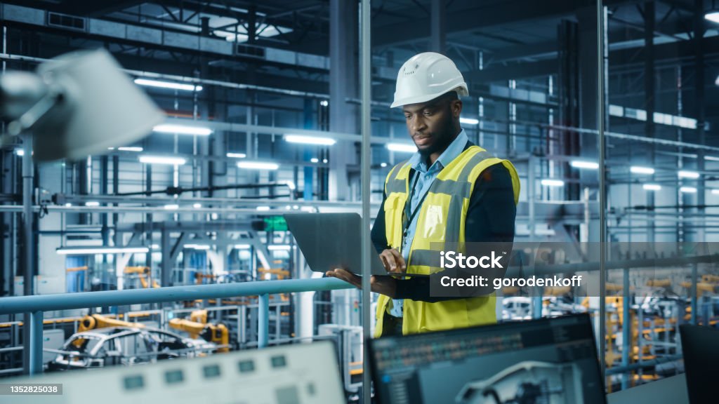 Car Factory: Professional Male Automotive Engineer Wearing Hard Hat, Walking, Using Laptop. Monitoring, Control, Equipment Production. Automated Robot Arm Assembly Line Manufacturing Electric Vehicles Manufacturing Stock Photo